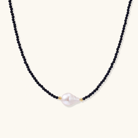 Delicate necklace with black blue sandstone beads and baroque pearl center on white background with gold accents.